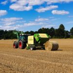 A tractor operates in a wheat field in Hohenhorn, harvesting hay bales on a sunny day.